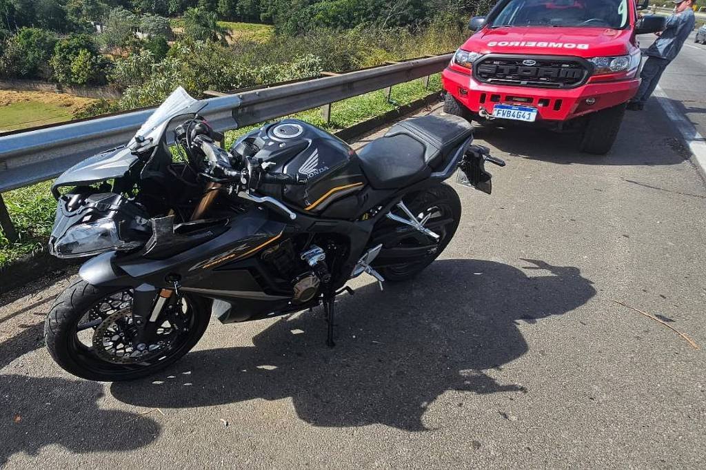 A imagem mostra uma motocicleta preta e dourada parada ao lado de uma estrada. A motocicleta está posicionada perto de uma barreira de proteção, com um fundo de vegetação e uma área rural visível ao longe. Ao fundo, há um veículo de apoio vermelho estacionado, que parece ser uma caminhonete. A estrada é pavimentada e há uma faixa de tráfego visível.