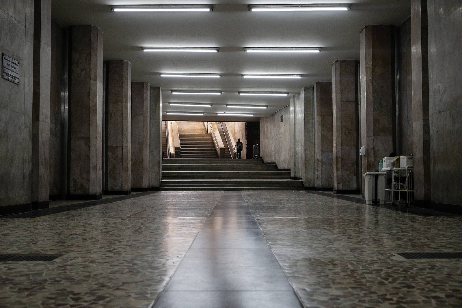 A imagem mostra um corredor amplo e vazio, com paredes de mármore e piso de ladrilhos. O teto é iluminado por luzes fluorescentes retangulares. Ao fundo, há uma escadaria que leva a um nível superior, e uma pessoa está visível perto das escadas. O ambiente parece ser de um edifício público ou comercial.
