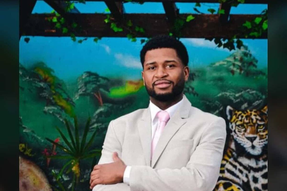 Um homem negro está posando em frente a um mural colorido que apresenta uma onça e vegetação tropical. Ele está vestido com um terno claro e uma gravata rosa, com os braços cruzados e um sorriso confiante. O fundo é vibrante, com tons de verde e detalhes da fauna.
