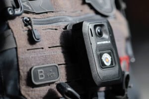 A imagem mostra uma câmera corporal da Motorola fixada em um colete tático. O colete é de cor cinza amarronzado e possui vários bolsos e detalhes em preto. A câmera é de formato retangular e possui um botão na parte frontal, além de um emblema visível. O fundo da imagem é desfocado, destacando o equipamento.