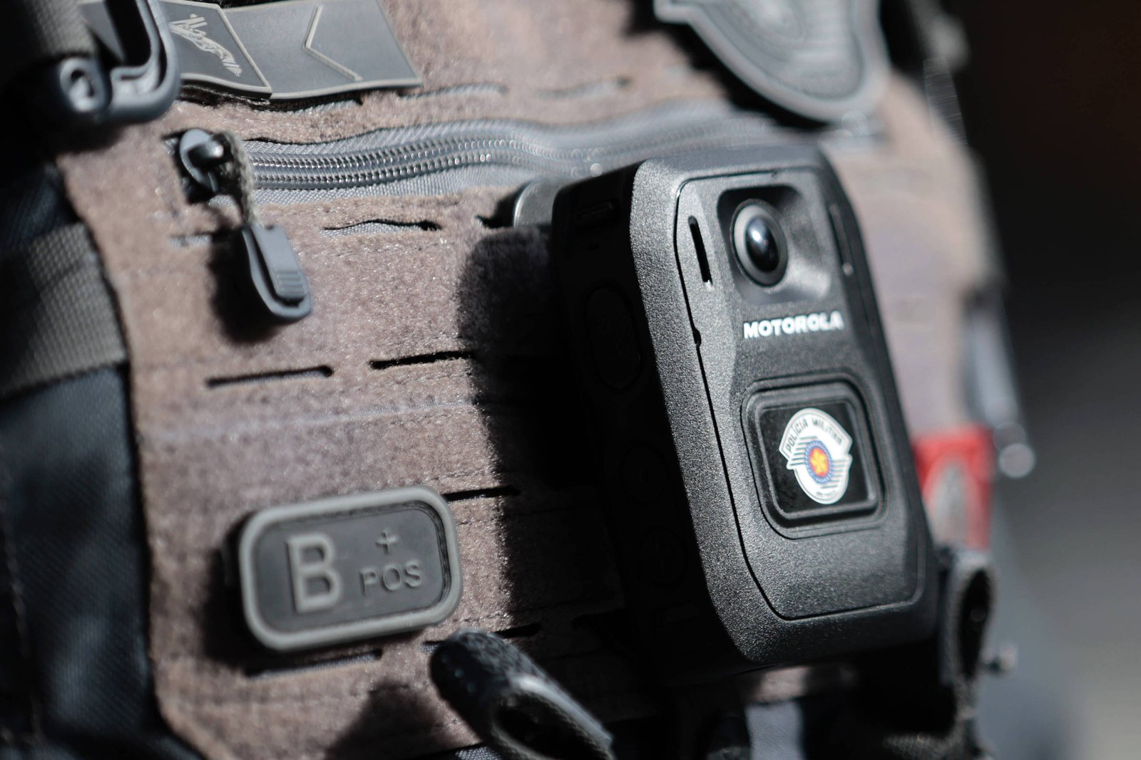 A imagem mostra uma câmera corporal da Motorola fixada em um colete tático. O colete é de cor cinza amarronzado e possui vários bolsos e detalhes em preto. A câmera é de formato retangular e possui um botão na parte frontal, além de um emblema visível. O fundo da imagem é desfocado, destacando o equipamento.