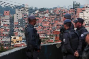 A imagem mostra um grupo de policiais em primeiro plano, vestidos com uniformes táticos e coletes à prova de balas, observando uma área urbana ao fundo. O cenário é composto por uma grande quantidade de prédios de apartamentos, predominantemente de cor vermelha e amarela, que se estendem em uma encosta. O céu está nublado, e há fios elétricos visíveis na parte superior da imagem.