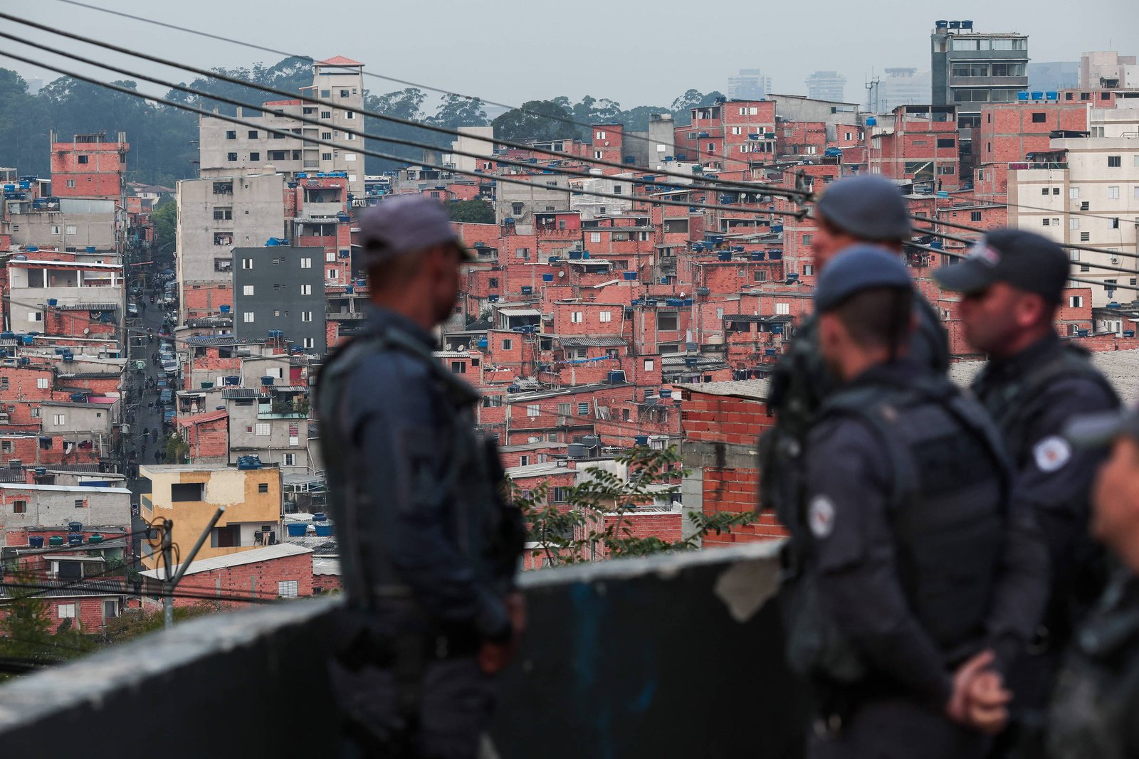 A imagem mostra um grupo de policiais em primeiro plano, vestidos com uniformes táticos e coletes à prova de balas, observando uma área urbana ao fundo. O cenário é composto por uma grande quantidade de prédios de apartamentos, predominantemente de cor vermelha e amarela, que se estendem em uma encosta. O céu está nublado, e há fios elétricos visíveis na parte superior da imagem.