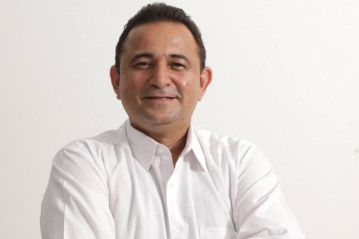 Um homem sorridente está posando para a foto, com os braços cruzados sobre uma cadeira azul. Ele usa uma camisa branca e tem cabelo curto e escuro. O fundo da imagem é claro e neutro, destacando o sujeito.