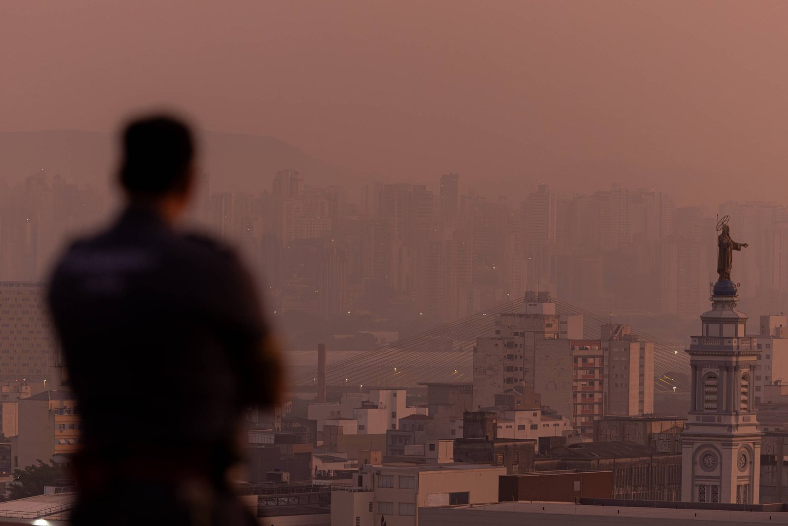 A imagem mostra uma vista panorâmica de uma cidade ao entardecer, com uma névoa que cobre os edifícios. Em primeiro plano, a silhueta de um policial é visível, de costas, observando a cena. Ao fundo, destacam-se prédios altos e uma torre de igreja com uma estátua no topo, que se sobressai na paisagem urbana.