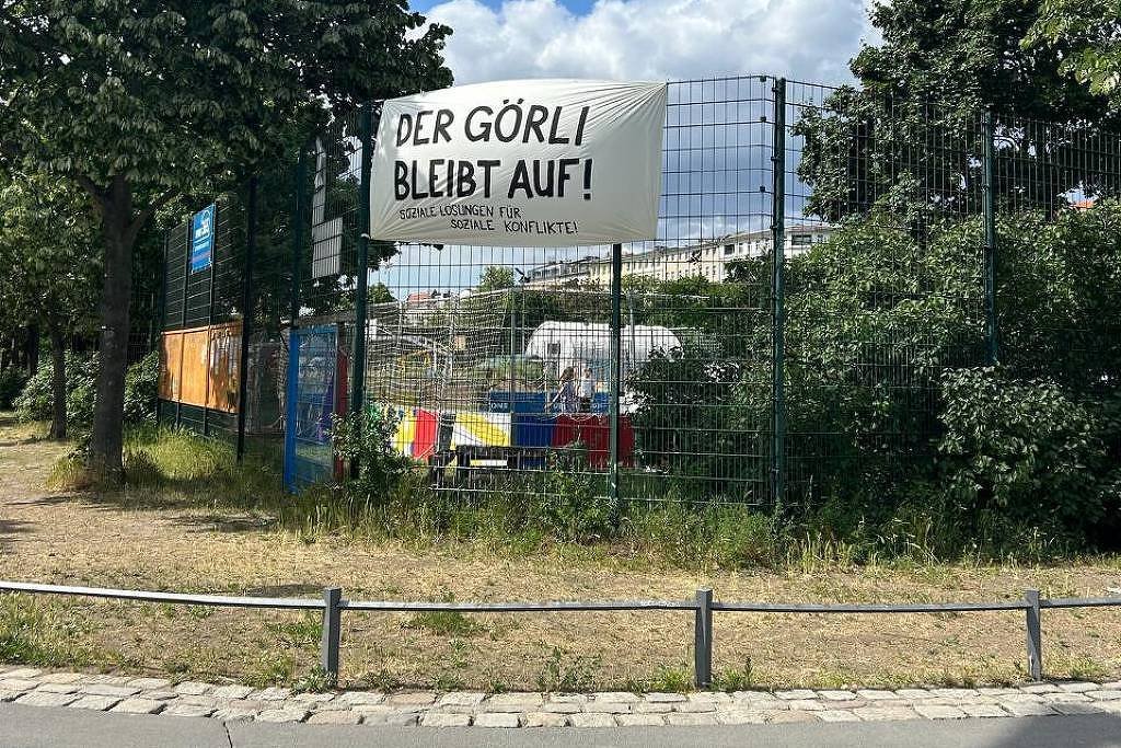 A imagem mostra uma faixa grande pendurada em uma cerca, com o texto