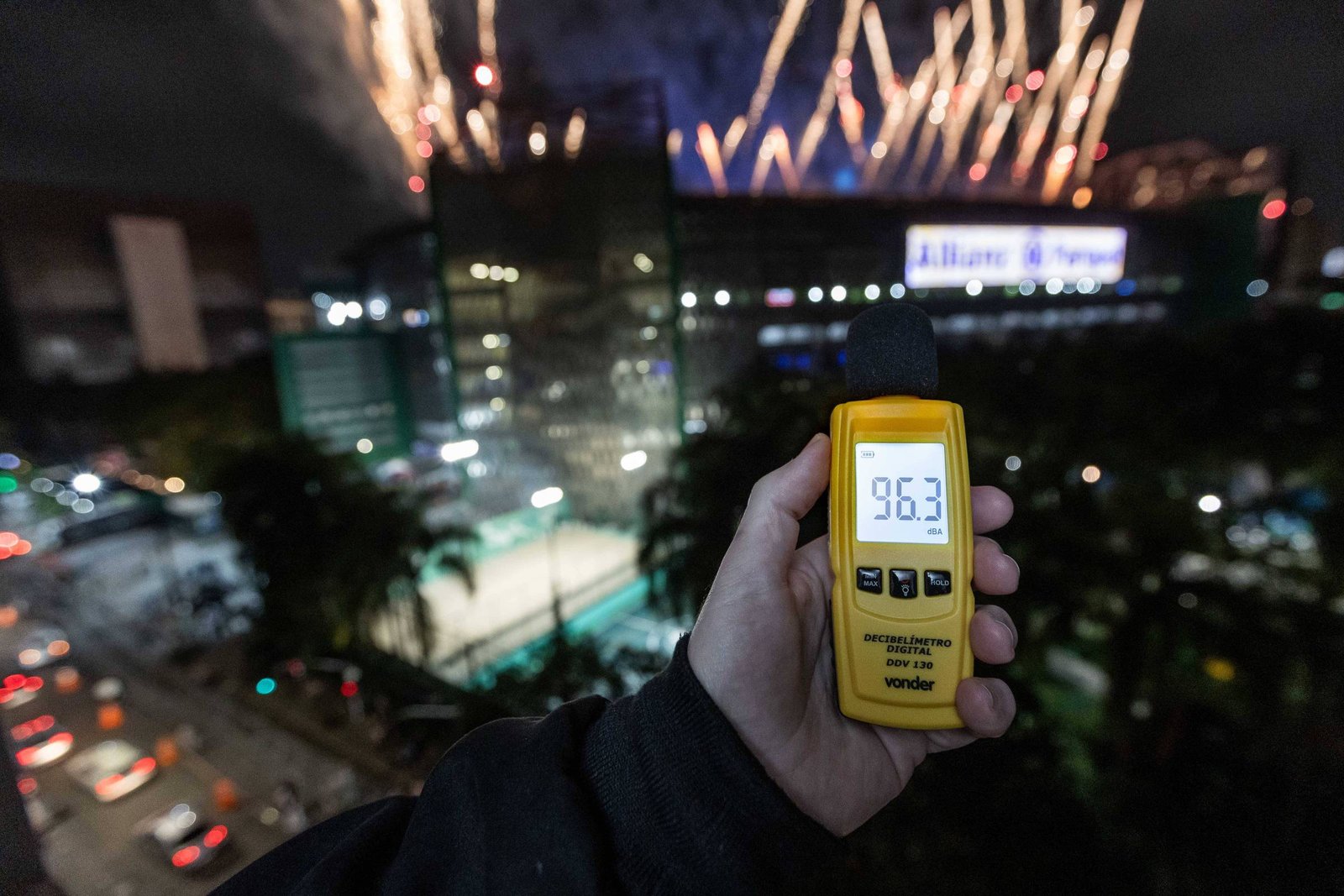 A imagem mostra uma mão segurando um medidor de decibéis, que exibe o valor de 96.3 dB. Ao fundo, há um espetáculo de fogos de artifício iluminando o céu noturno, com um grande edifício visível, possivelmente um estádio, onde está escrito