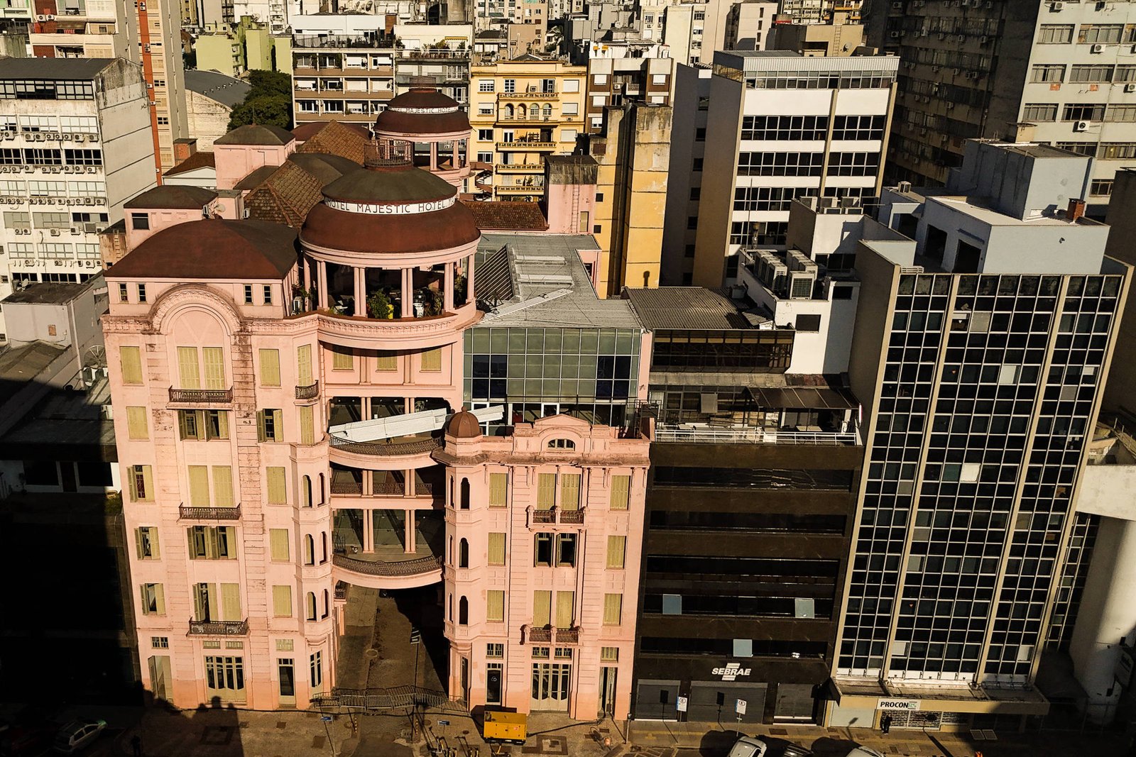 A imagem mostra um edifício rosa de arquitetura distinta, localizado em uma área urbana densa. O prédio possui várias janelas e um design que se destaca entre os edifícios ao redor, que são predominantemente de cores neutras e modernas. O entorno é composto por prédios altos e uma vista da cidade ao fundo, com um céu claro.