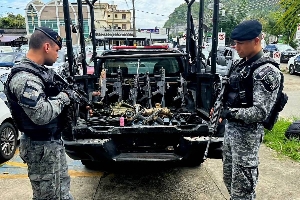 Dois policiais em uniforme tático estão em frente a uma caminhonete. A caçamba da caminhonete está carregada com várias armas, incluindo fuzis. Os policiais estão observando as armas, um deles segura um fuzil e o outro está com uma arma em mãos. Ao fundo, há outros veículos estacionados e uma área urbana com prédios e montanhas ao longe.