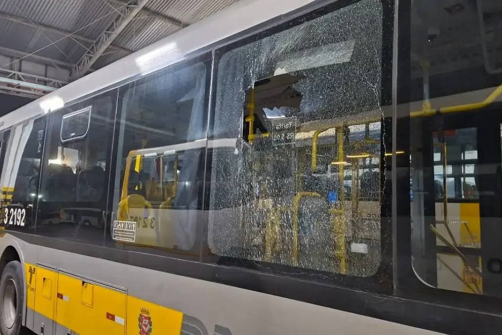 A imagem mostra um ônibus estacionado em um local coberto, com um vidro lateral quebrado. O vidro está estilhaçado, e a parte interna do ônibus é visível, com assentos e outros elementos do interior. O ônibus é de cor amarela e cinza, e há um número de identificação visível na lateral, que é 2102.