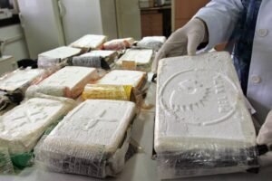 A imagem mostra uma mesa com vários pacotes embrulhados em plástico, que parecem conter substâncias ilícitas. Um dos pacotes está sendo segurado por uma pessoa com luvas brancas, e possui uma marca visível. Ao fundo, há uma cozinha ou laboratório com armários e um refrigerador.