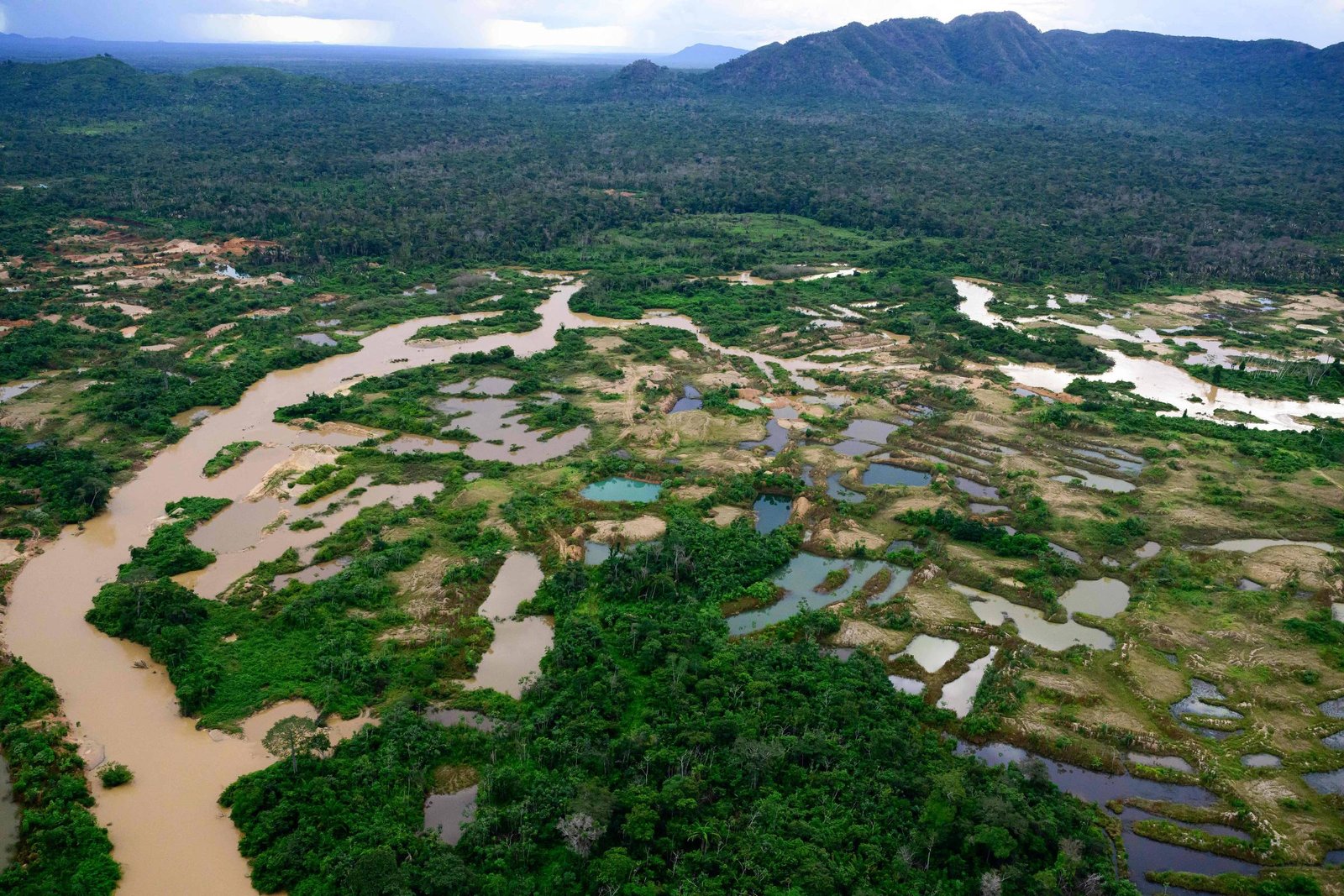A imagem mostra uma vista aérea de uma vasta área florestal com lagoas de garimpo. As lagoas têm águas de diferentes tonalidades, variando entre marrom e azul, e estão cercados por vegetação densa. Ao fundo, há montanhas cobertas de árvores, e o céu apresenta nuvens.