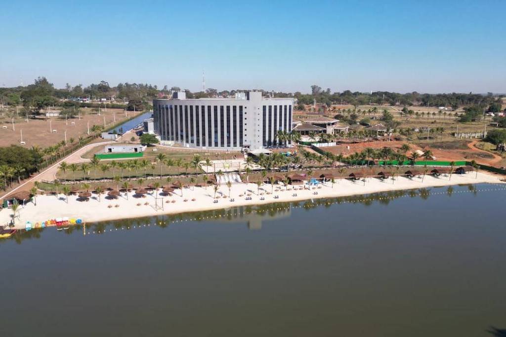 A imagem mostra um hotel moderno localizado à beira de um lago. O edifício é grande e possui várias janelas. Na frente do hotel, há uma praia de areia clara com espreguiçadeiras e guarda-sóis. O lago é calmo e reflete o céu azul. Ao fundo, há áreas verdes e outras construções, sugerindo um ambiente de lazer e relaxamento.