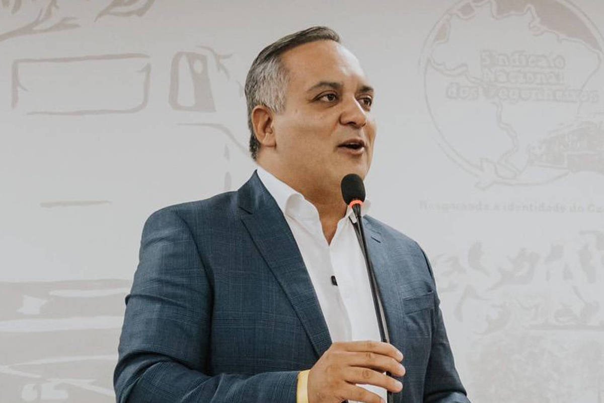 Um homem de terno azul e camisa branca está falando ao microfone em um evento. Ele parece estar em um palco ou em uma mesa de conferência, com um fundo que possui desenhos ou ilustrações. Ao fundo, há um outro homem sentado, observando. O ambiente parece ser formal, possivelmente uma reunião ou conferência.