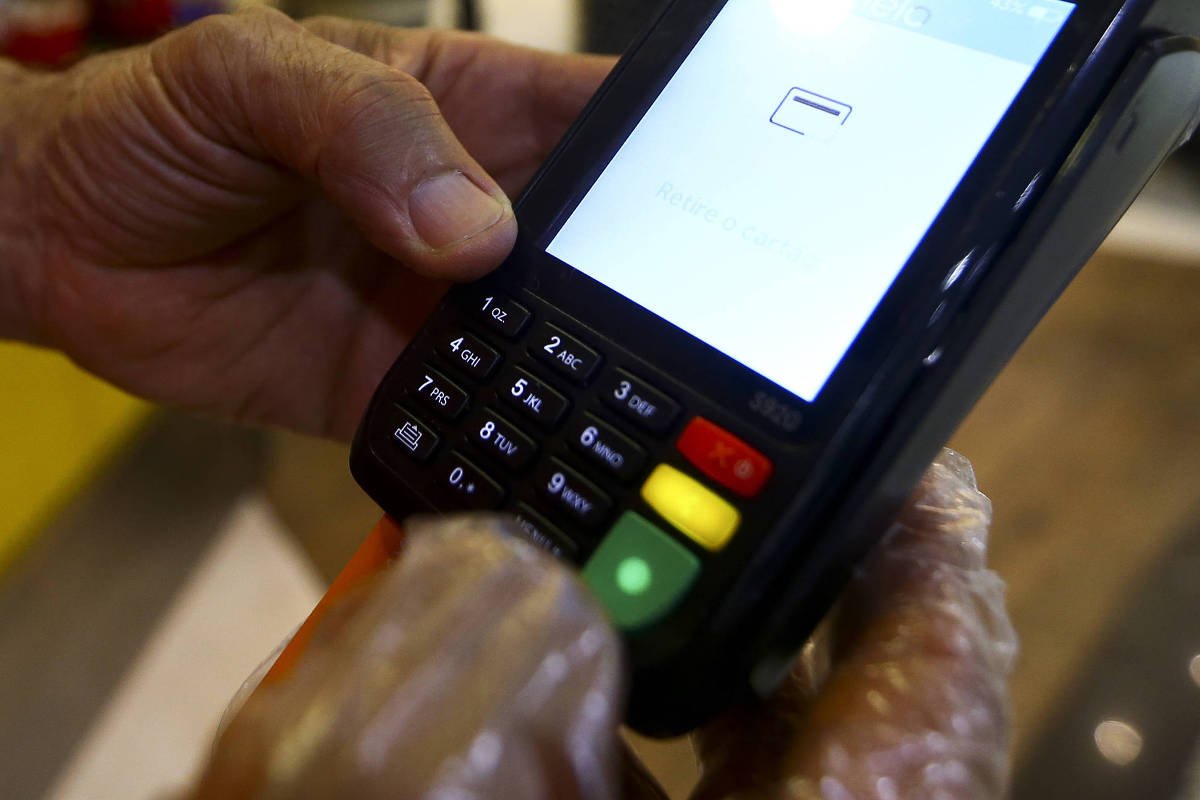 A imagem mostra uma mão segurando um terminal de pagamento eletrônico. O dispositivo possui uma tela que está exibindo um ícone de carregamento ou espera. Abaixo da tela, há um teclado com botões coloridos, incluindo um botão verde, um botão vermelho e um botão amarelo. A mão que segura o terminal está usando uma luva plástica.