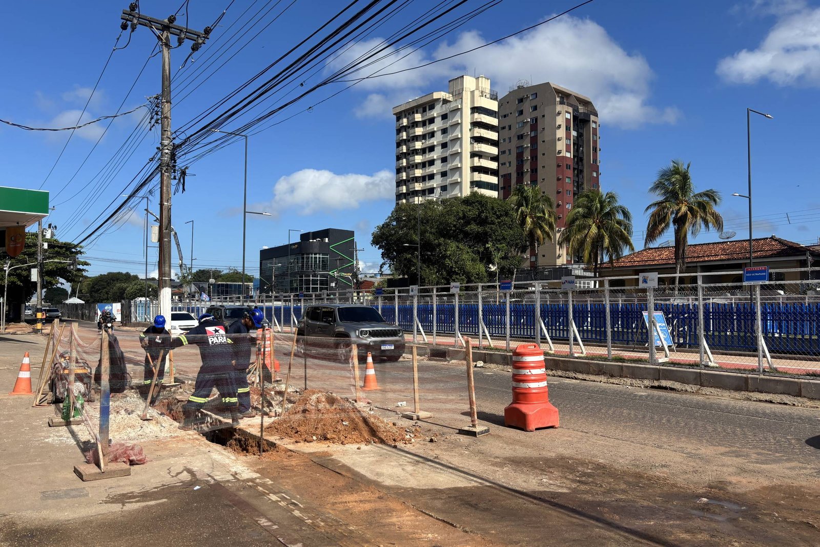 A imagem mostra uma rua em obras, com um buraco aberto no asfalto. Há cones laranjas e barreiras de segurança ao redor da área em construção. No fundo, é possível ver prédios altos e algumas palmeiras. O céu está claro e ensolarado, com algumas nuvens.