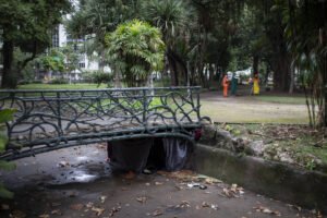 A imagem mostra uma ponte de ferro em um parque urbano. A ponte é de cor verde e está sobre um pequeno canal. Abaixo da ponte, há um espaço coberto por tecidos escuros e alguns detritos no chão. Ao fundo, é possível ver árvores e duas pessoas vestindo uniformes laranja e amarelo, que parecem estar realizando alguma atividade no parque.