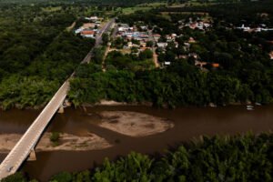A imagem mostra uma vista aérea de uma área urbana cercada por vegetação. À esquerda, há uma ponte que atravessa um rio de águas escuras. Abaixo da ponte, é visível uma pequena ilha de terra no meio do rio. À direita, há uma parte da cidade com casas e ruas, cercada por árvores e vegetação densa. O céu está limpo e a luz do sol ilumina a cena.
