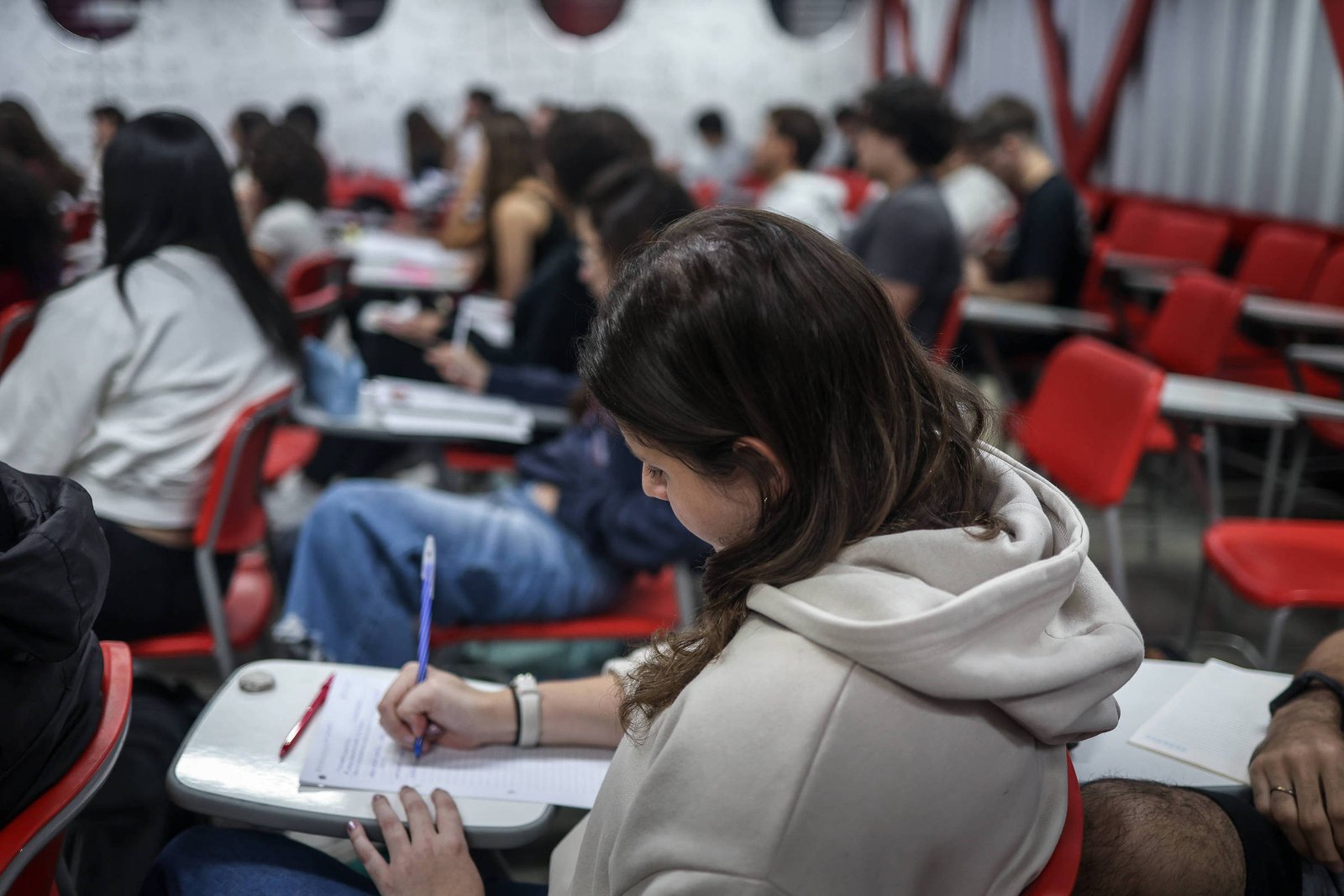 A imagem mostra uma sala de aula com vários alunos sentados em cadeiras vermelhas. No primeiro plano, uma estudante está escrevendo em um caderno, segurando uma caneta azul. Ao fundo, outros alunos estão visíveis, alguns de costas, em um ambiente com paredes brancas e detalhes em vermelho.