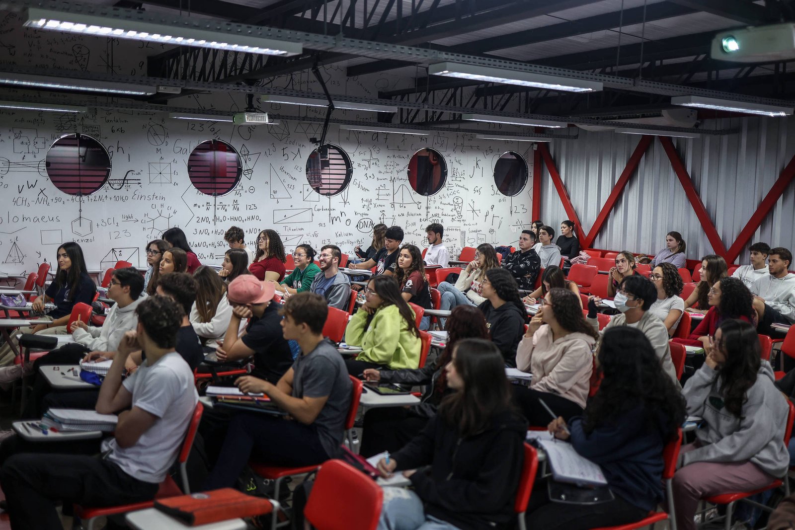 A imagem mostra uma sala de aula com muitos alunos sentados em cadeiras vermelhas. O ambiente é iluminado e as paredes estão cobertas com anotações e fórmulas matemáticas. Os alunos estão atentos, alguns olhando para frente e outros conversando. Há janelas redondas na parede, que permitem a entrada de luz natural.