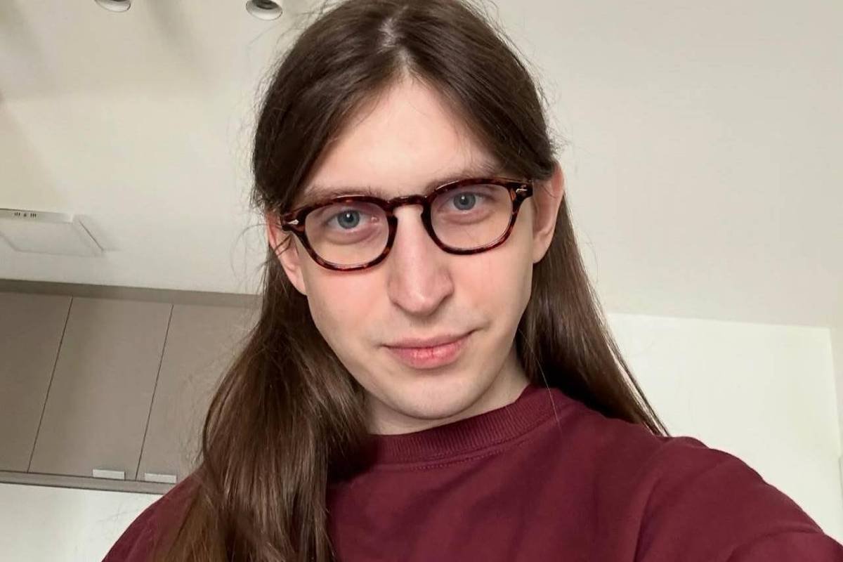 A imagem mostra uma pessoa com cabelo longo e liso, usando óculos e uma camiseta de manga longa na cor marrom. A pessoa está em um ambiente interno, com uma cozinha ao fundo. A iluminação é clara e suave, destacando o rosto da pessoa.