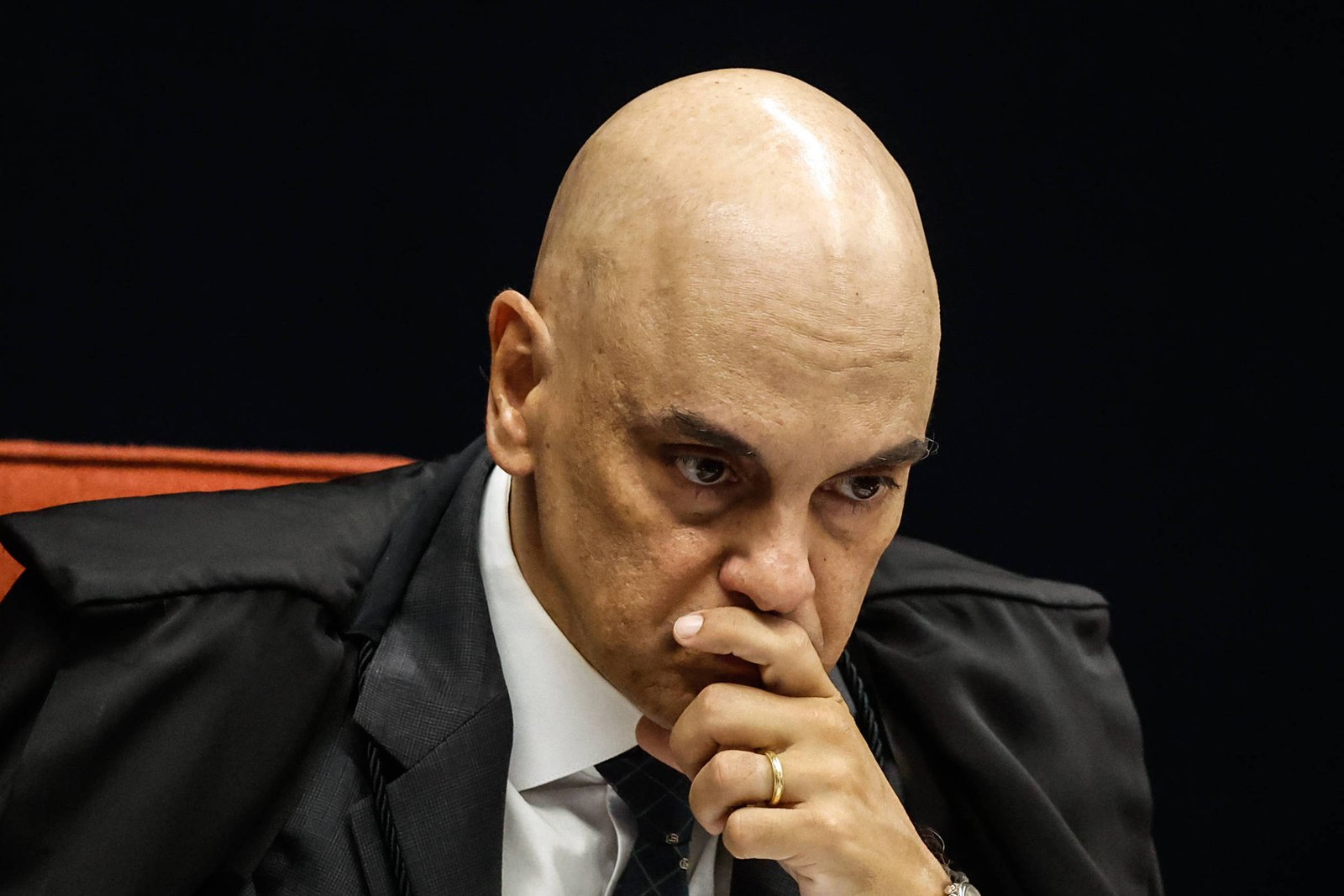 Um homem careca, vestido com uma toga preta, está sentado em uma cadeira vermelha. Ele parece pensativo, com a mão cobrindo parcialmente o rosto e os olhos voltados para baixo. O fundo é escuro, destacando sua expressão séria e contemplativa.