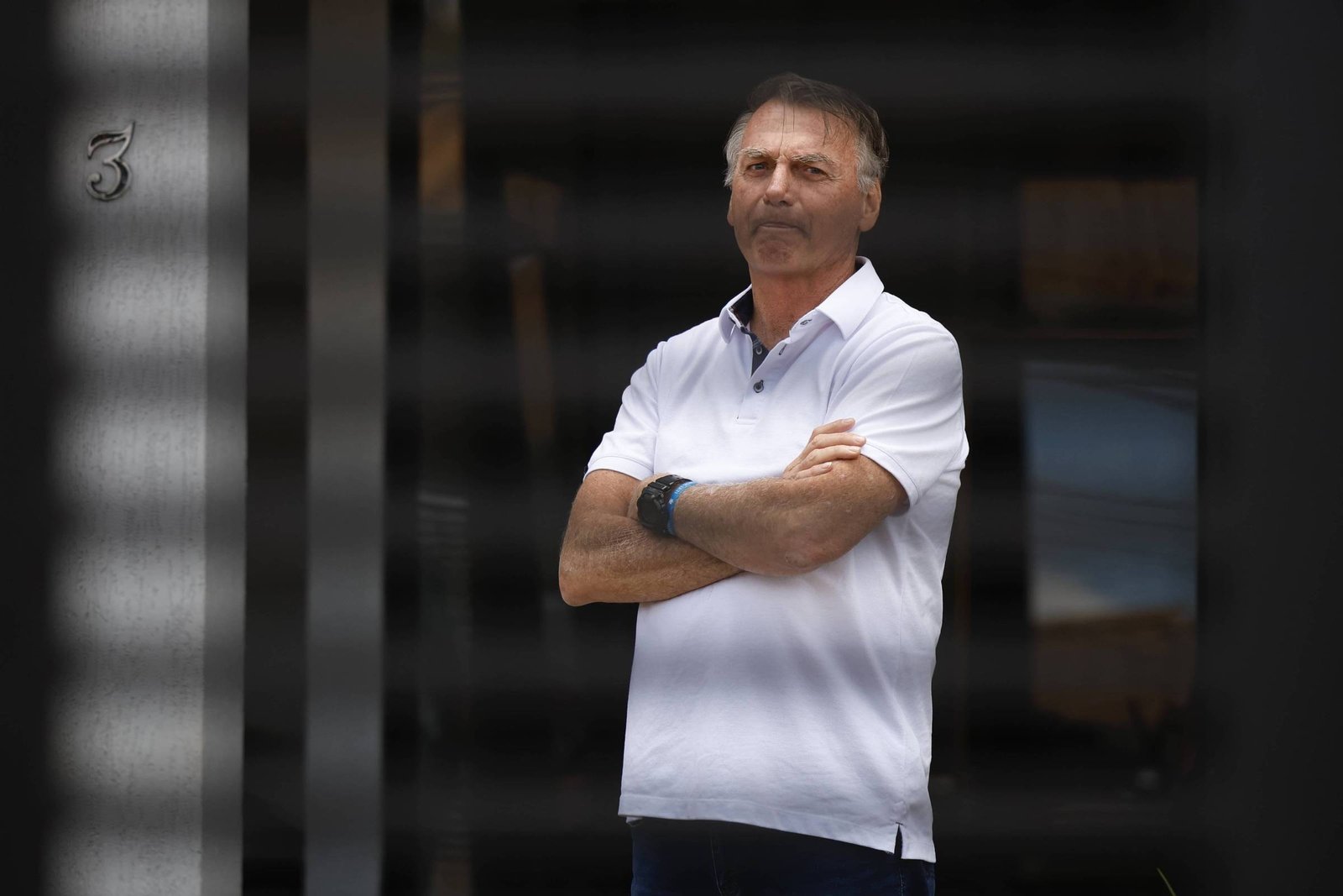 Um homem está em pé com os braços cruzados, usando uma camisa polo branca e calças escuras. Ele tem cabelo curto e está olhando para a frente. Ao fundo, há uma parede com uma porta e o número 3 visível. A imagem tem um efeito de grade, que obscurece parcialmente a visão do homem.