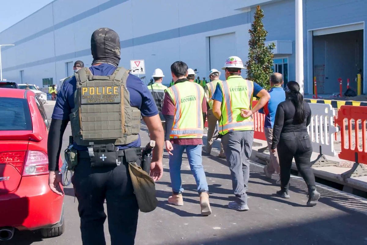 A imagem mostra um grupo de pessoas em uma área industrial. À frente, um policial com colete tático e máscara caminha em direção a um grupo de trabalhadores que usam coletes refletivos. Ao fundo, há um prédio industrial e veículos estacionados. A cena parece ser parte de uma operação de fiscalização ou segurança.