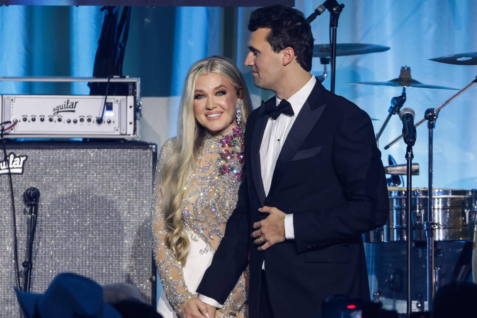 Um casal está em um palco durante um evento. A mulher, com cabelo longo e liso, usa um vestido brilhante e está sorrindo. O homem, ao seu lado, veste um terno preto e uma camisa branca. Ao fundo, há equipamentos de som, incluindo amplificadores e instrumentos musicais.