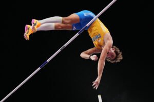 Um atleta está realizando um salto com vara, elevado acima de uma barra horizontal. Ele usa uma camiseta amarela e calças azuis, com meias brancas e tênis coloridos. O fundo é escuro, destacando a ação do salto.