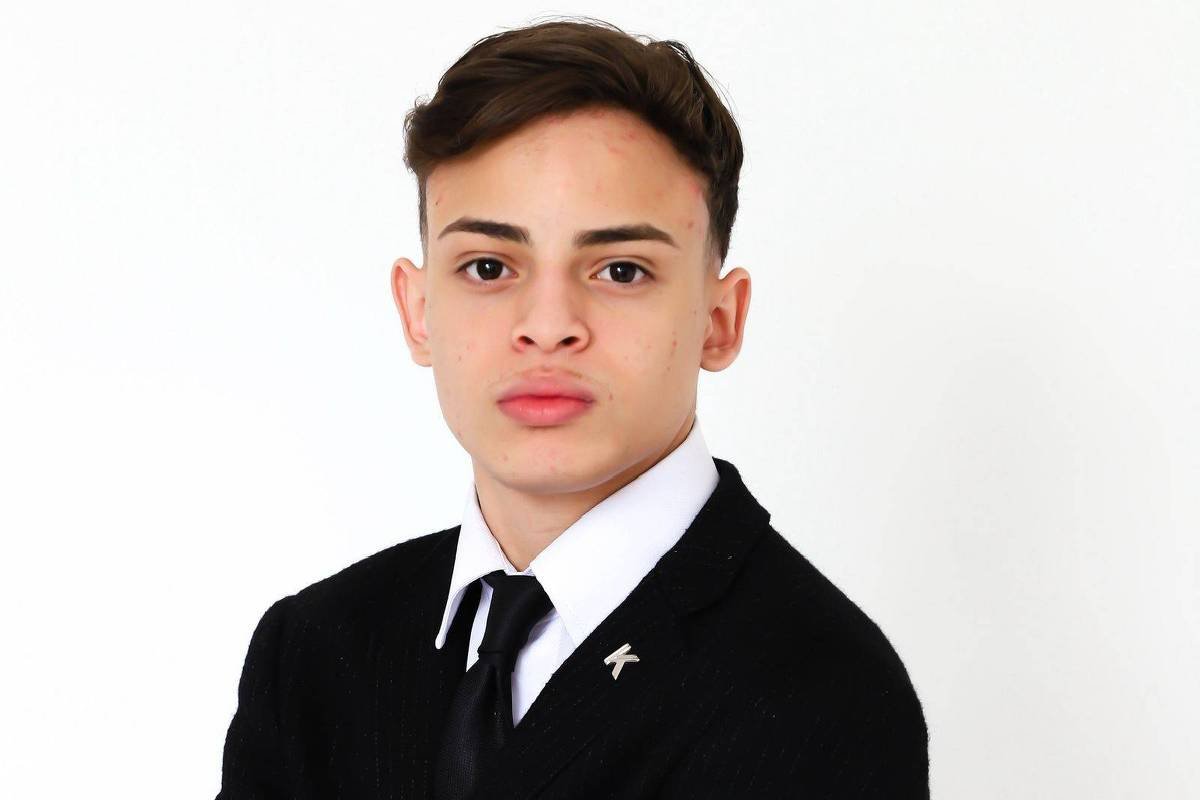 Um jovem está posando em um fundo branco, vestindo um terno preto com uma camisa branca e uma gravata preta. Ele tem os braços cruzados e uma expressão séria no rosto. O cabelo é curto e bem arrumado, e há um pequeno broche em forma de