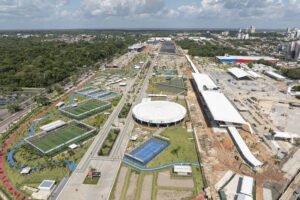 A imagem mostra uma vista aérea de uma área esportiva em construção, com campos de futebol e quadras de tênis visíveis. Há um grande estádio circular no centro da imagem, cercado por áreas verdes e estruturas em construção. Ao fundo, pode-se ver uma cidade com prédios e uma vegetação densa ao redor. O céu está parcialmente nublado.