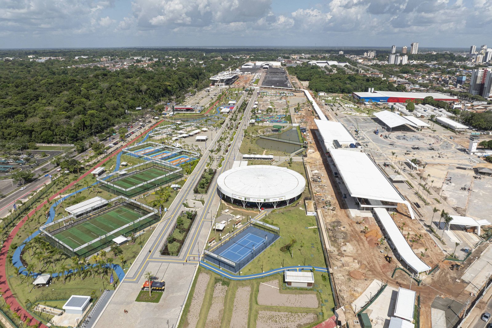 A imagem mostra uma vista aérea de uma área esportiva em construção, com campos de futebol e quadras de tênis visíveis. Há um grande estádio circular no centro da imagem, cercado por áreas verdes e estruturas em construção. Ao fundo, pode-se ver uma cidade com prédios e uma vegetação densa ao redor. O céu está parcialmente nublado.