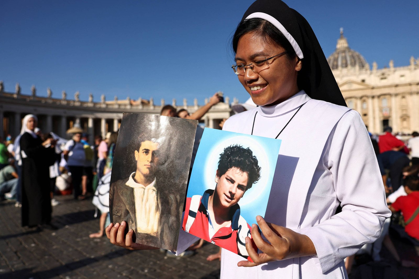 Uma freira sorridente está em pé em uma praça, segurando duas fotos. A foto da esquerda mostra um homem com um retrato em preto e branco, enquanto a da direita apresenta um jovem com cabelo escuro e uma camiseta vermelha. Ao fundo, há uma multidão de pessoas e a arquitetura de um edifício, possivelmente uma basílica, sob um céu azul claro.