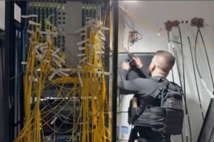 A imagem mostra um homem com um colete tático, de costas, em uma sala de servidores. Ele está próximo a um rack de servidores que possui muitos cabos amarelos desorganizados. O homem parece estar ajustando ou inspecionando um equipamento na parede ao lado do rack. O ambiente é iluminado artificialmente e há outros cabos visíveis na parte superior da imagem.