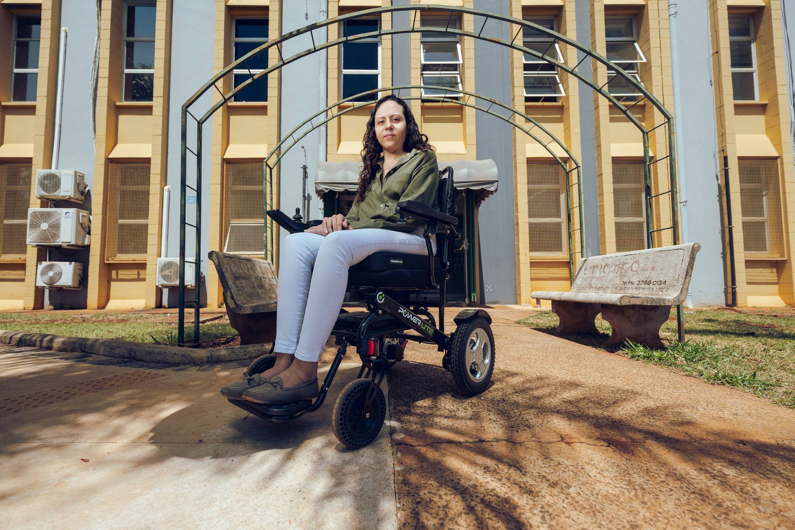Mulher jovem sentada em cadeira de rodas motorizada no caminho de concreto diante de um prédio com janelas altas e ar-condicionado externo. Estrutura metálica arqueada está atrás dela, e banco de concreto está à direita.