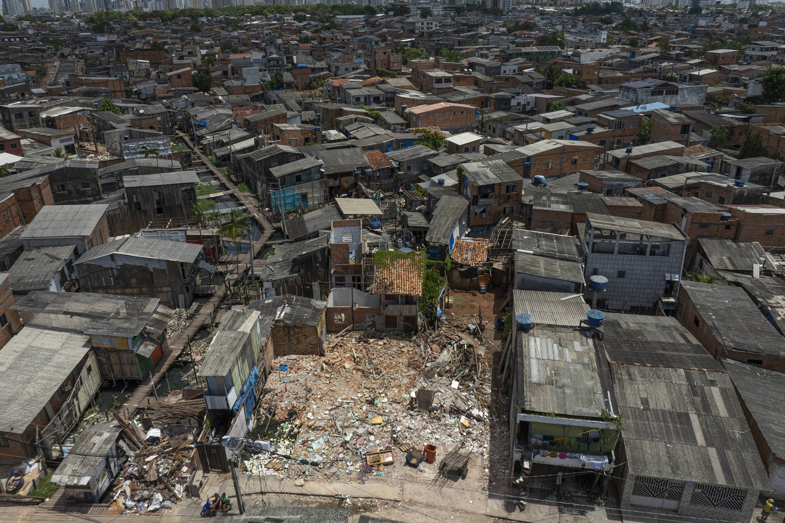 A imagem mostra uma vista aérea de uma comunidade urbana, caracterizada por uma densa aglomeração de casas de construção simples e precária. Algumas casas estão em estado de deterioração, e há áreas com entulho e lixo visíveis. Ao fundo, é possível ver um horizonte urbano com prédios altos, contrastando com a área mais baixa da comunidade.