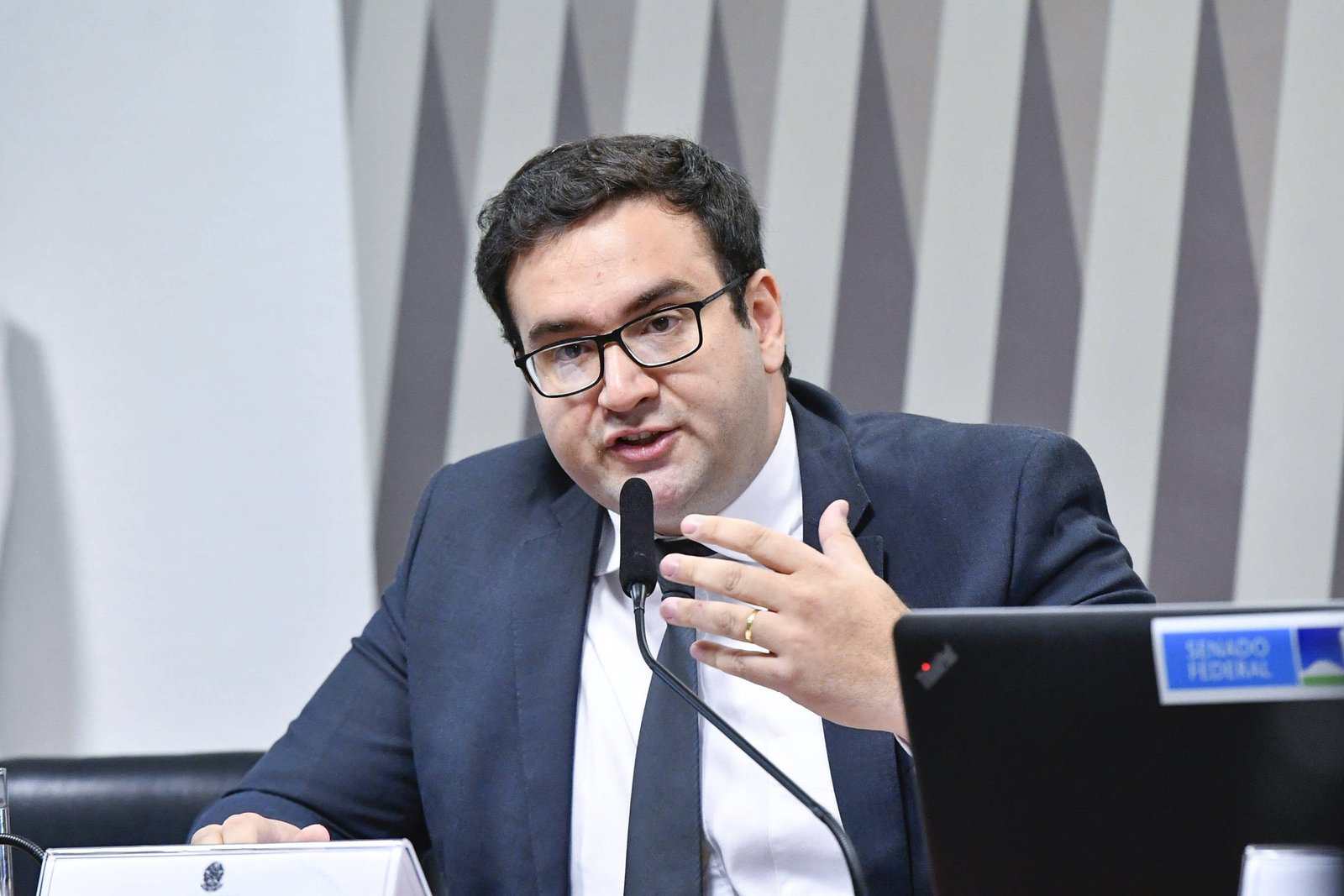 Um homem com óculos e terno escuro está falando em um microfone durante uma audiência no Senado. Ele gesticula com a mão direita enquanto se dirige a um público. Ao fundo, há uma mesa com um laptop e uma placa que diz