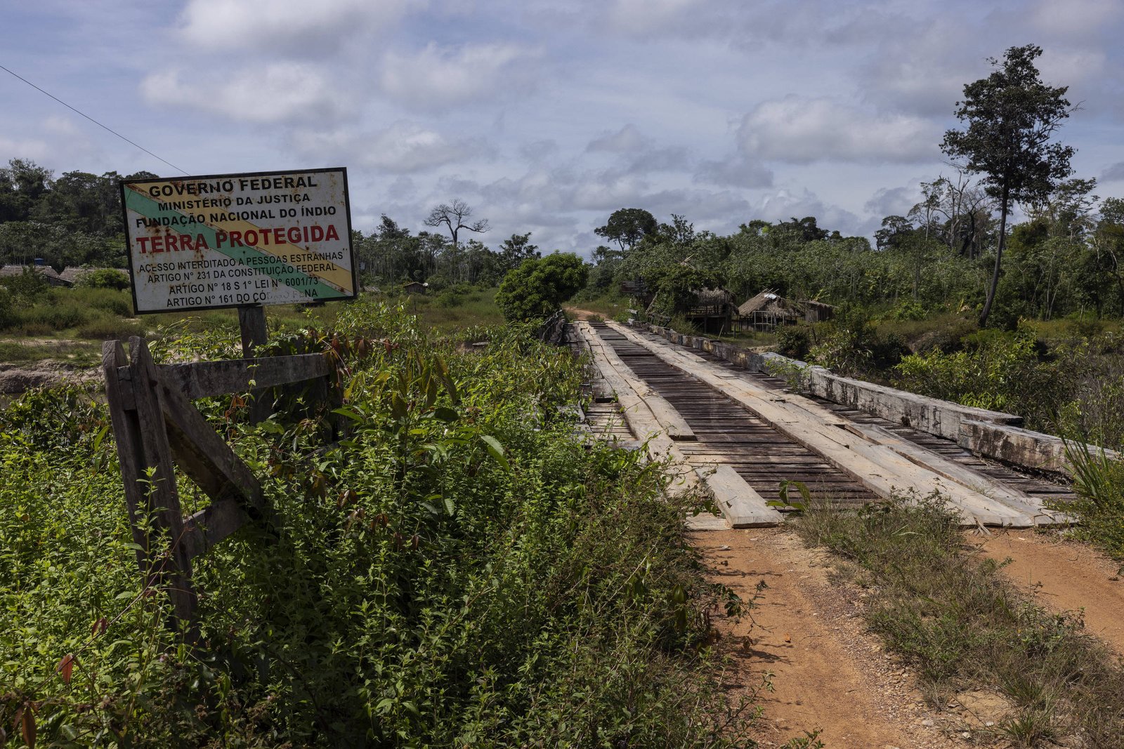 A imagem mostra uma ponte de madeira em uma área rural, cercada por vegetação densa. À esquerda, há uma placa do Governo Federal do Brasil, indicando que a área é uma Terra Protegida. O céu está parcialmente nublado, e a estrada de terra leva em direção à ponte.