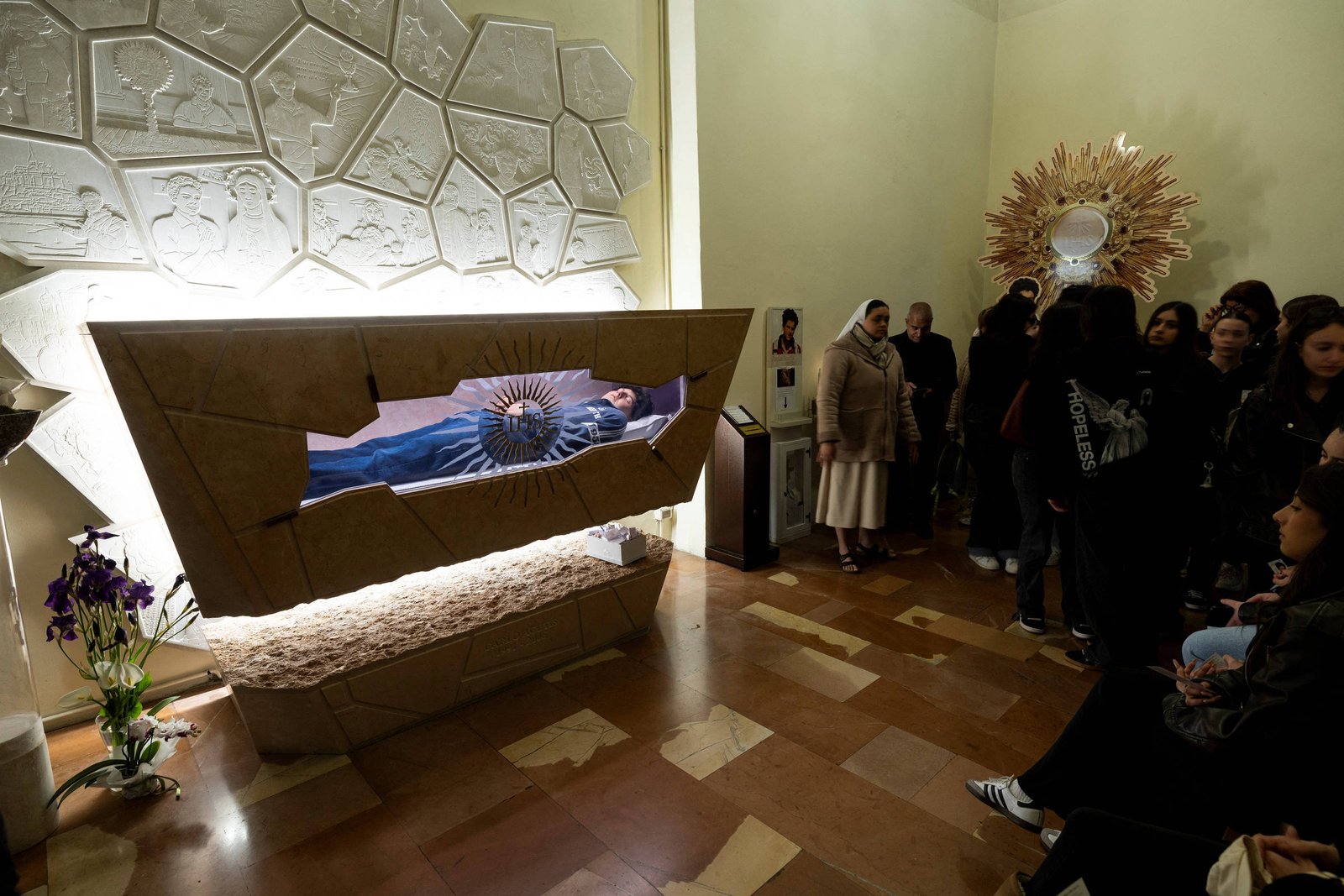 A imagem mostra uma exposição em um espaço interno, onde há um sarcófago estilizado com uma figura deitado dentro, coberta por um manto azul. O sarcófago é cercado por uma estrutura de pedra com relevos e há flores roxas na frente. Um grupo de pessoas está em pé ao lado, observando a obra, enquanto uma mulher vestida de hábito religioso está à esquerda. O ambiente é iluminado e possui um piso de ladrilhos.