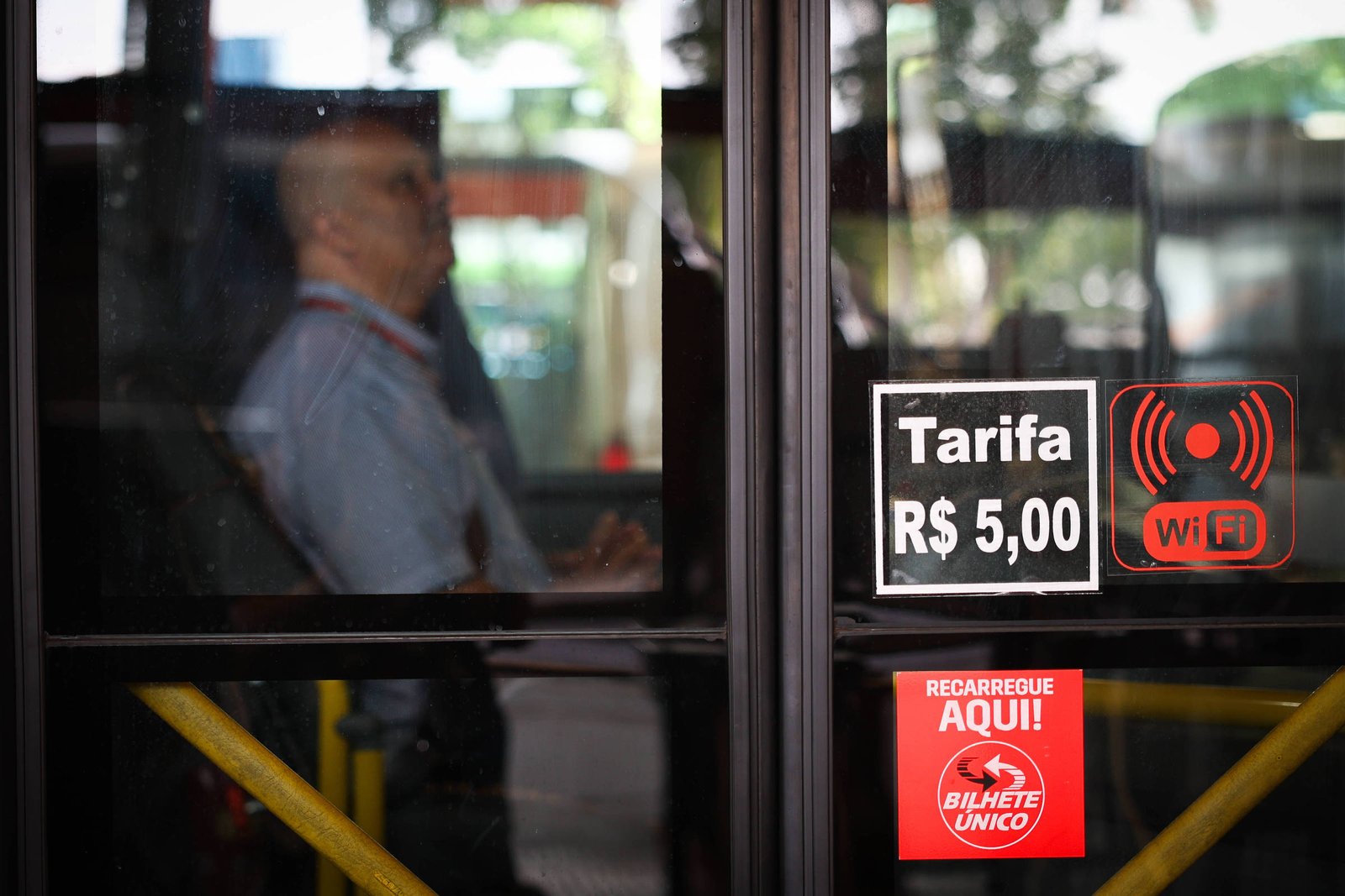 Passageiro sentado dentro de ônibus visto através da porta de vidro. Na porta, adesivos indicam tarifa de R$ 5,00, disponibilidade de Wi-Fi e ponto para recarga do Bilhete Único.