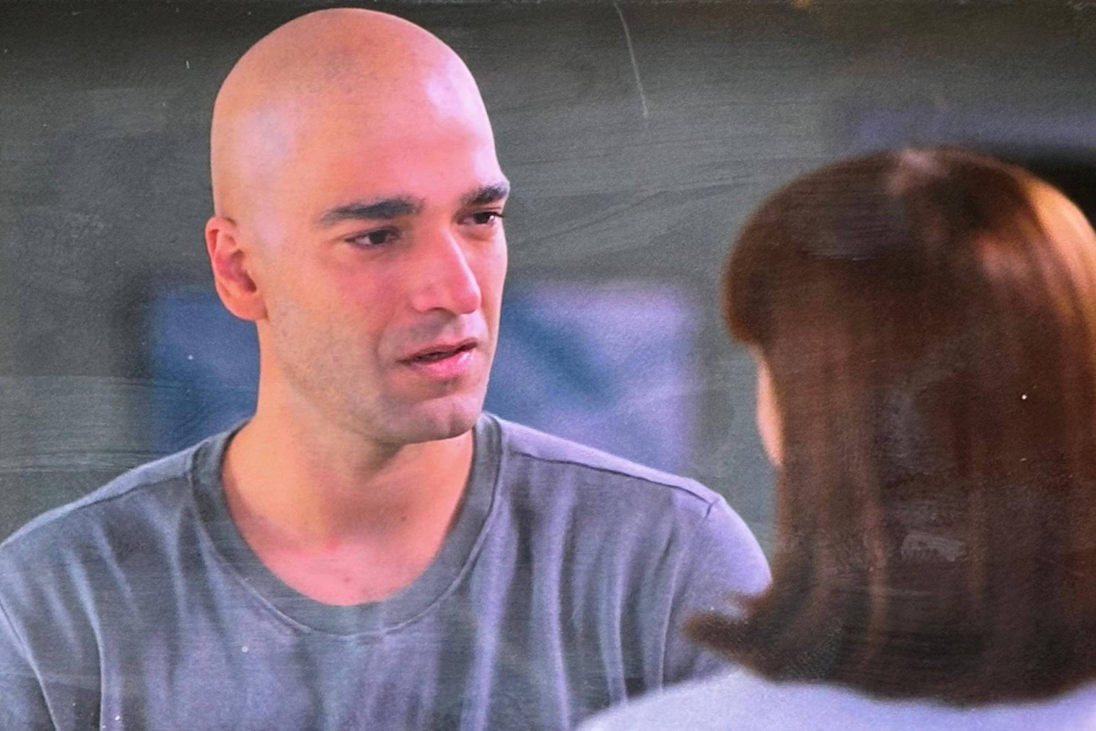 Um homem careca, vestindo uma camiseta cinza, está conversando com uma mulher de cabelo castanho. O homem parece estar emocionalmente envolvido na conversa, enquanto a mulher escuta atentamente. O fundo é desfocado, sugerindo um ambiente interno.