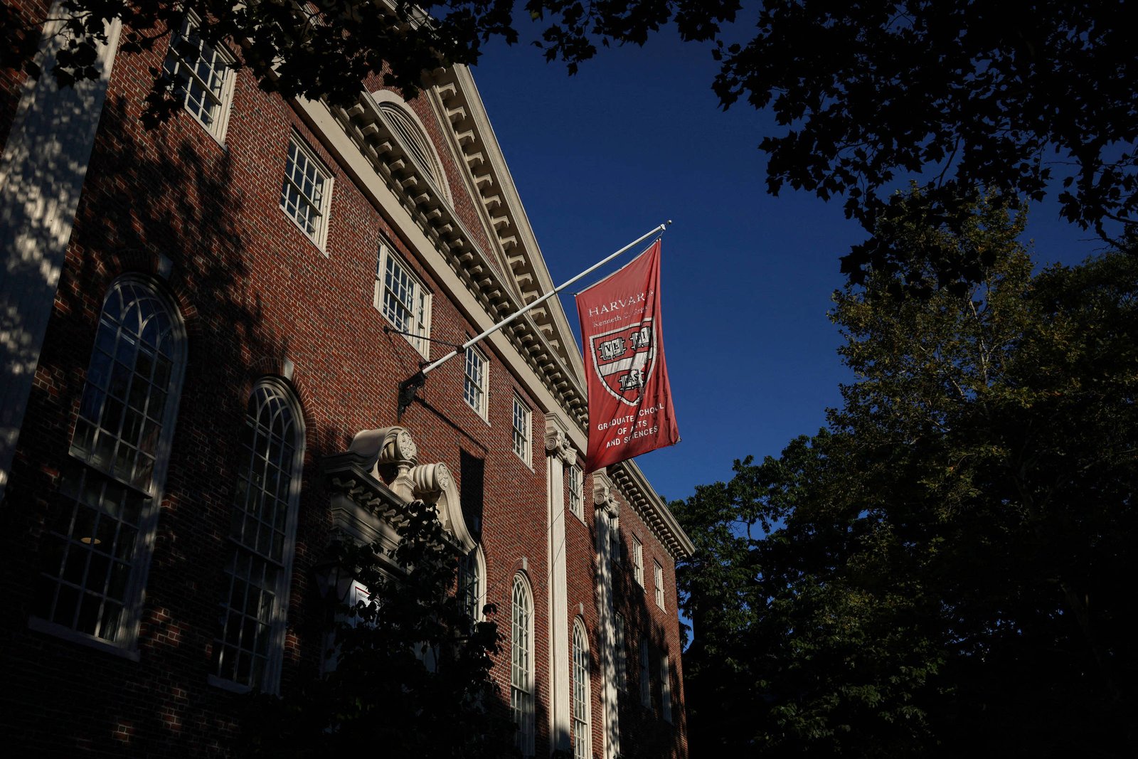 Prédio de tijolos vermelhos com janelas arqueadas e bandeira vermelha de Harvard pendurada em mastro, sob céu azul claro e árvores ao redor.