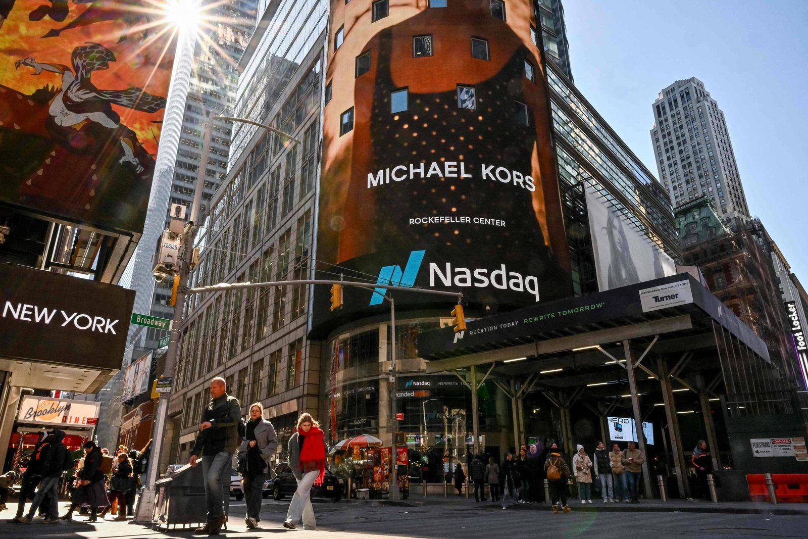 Painel eletrônico grande exibe anúncio da Michael Kors no Rockefeller Center, com logo da Nasdaq abaixo. Pessoas caminham na calçada sob luz solar intensa, cercadas por prédios altos no centro de Nova York.