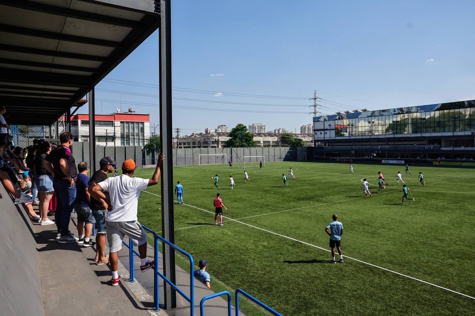 Público acompanha partida de futebol em arquibancada coberta ao lado de campo gramado. Jogadores em uniformes brancos e verdes disputam bola sob céu azul, com edifícios ao fundo.