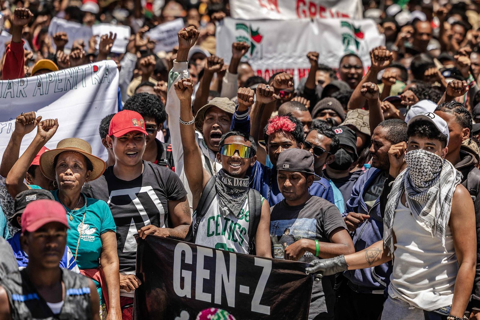 Grupo numeroso de jovens em manifestação ao ar livre, muitos com punhos erguidos. Alguns seguram faixa preta com texto