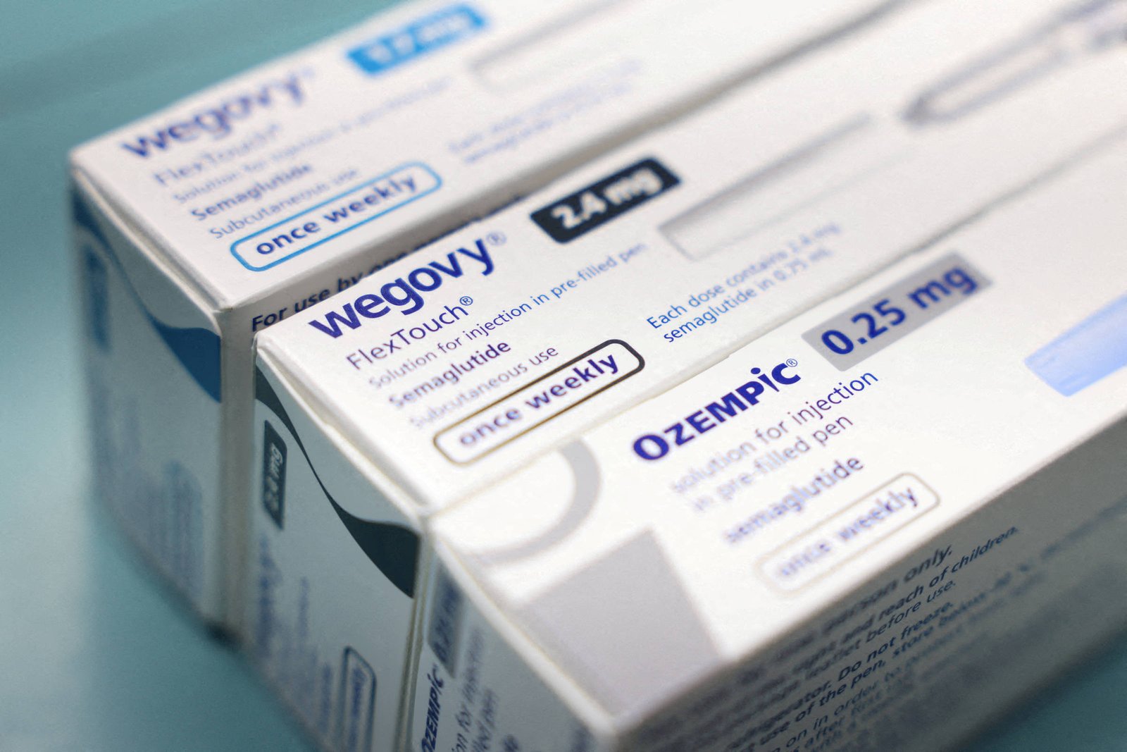 Duas caixas de medicamentos Wegovy e Ozempic posicionadas lado a lado sobre superfície azul. Ambas indicam uso semanal e contêm a substância semaglutida, com dosagens de 1,2 mg e 0,25 mg visíveis nas embalagens.