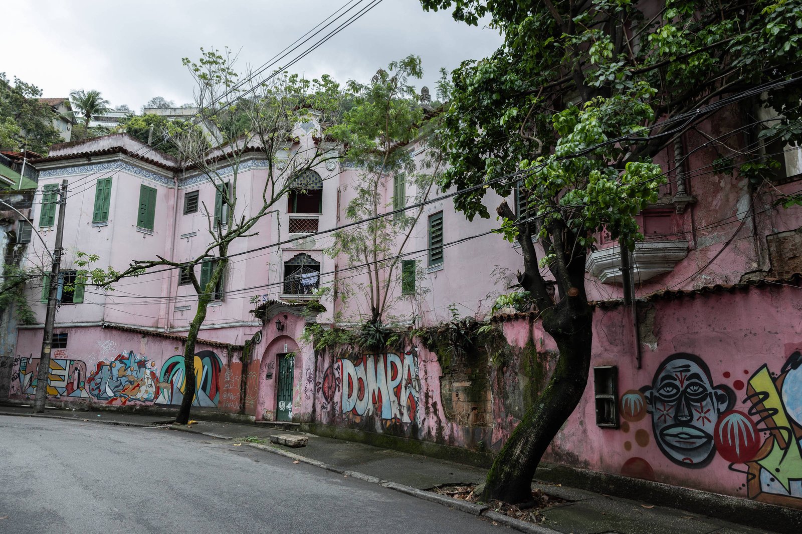 Prédio antigo de dois andares com paredes rosas e janelas verdes, localizado em rua curva. A fachada está coberta por grafites coloridos e árvores com galhos que se estendem sobre a calçada e a rua. O céu está nublado.