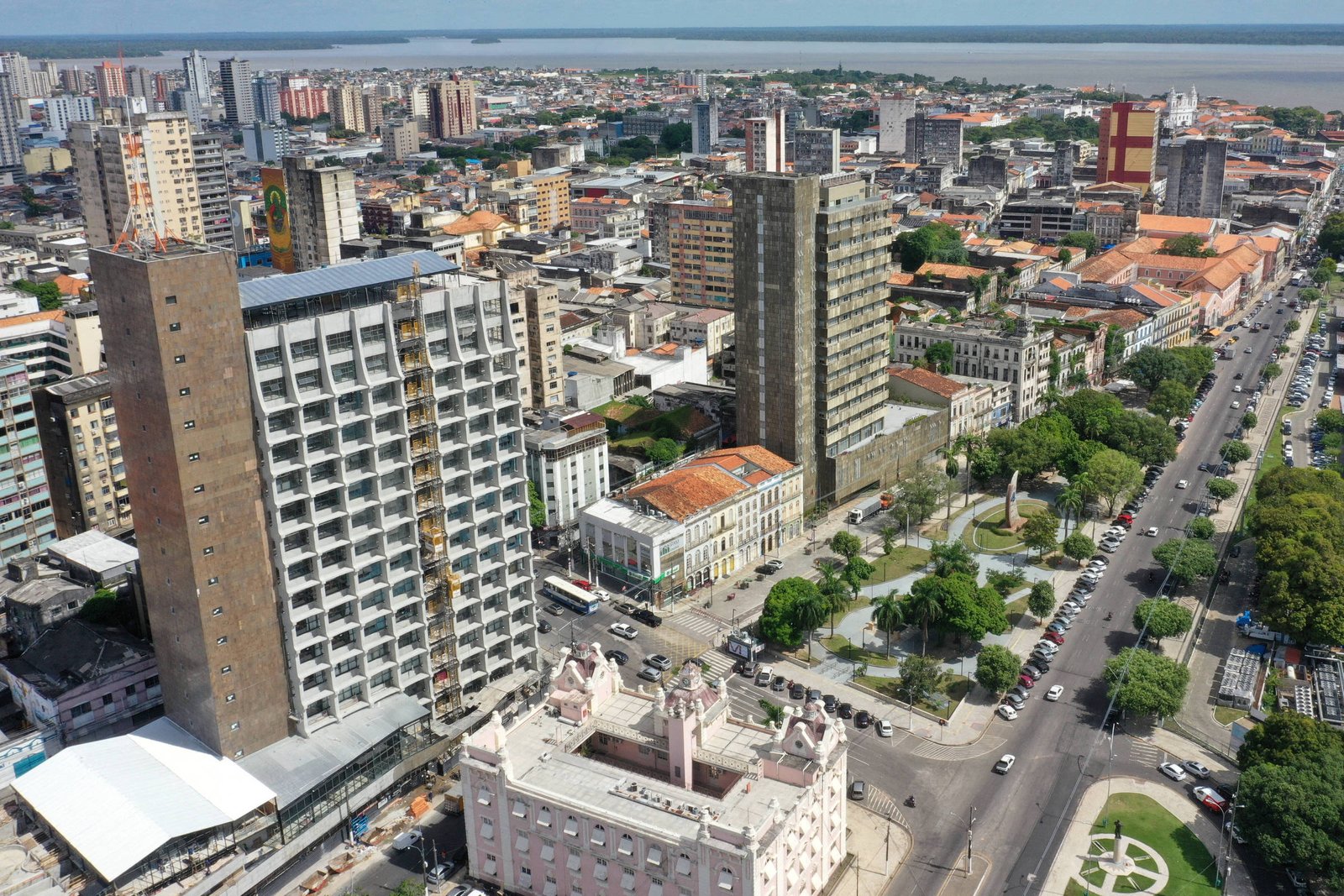 A imagem mostra uma vista aérea de uma cidade com vários edifícios altos e modernos. No primeiro plano, há um edifício de vários andares com uma fachada de vidro e concreto. Ao fundo, é possível ver uma mistura de construções, incluindo prédios residenciais e comerciais, além de áreas verdes. A cidade parece estar localizada próxima a um corpo d