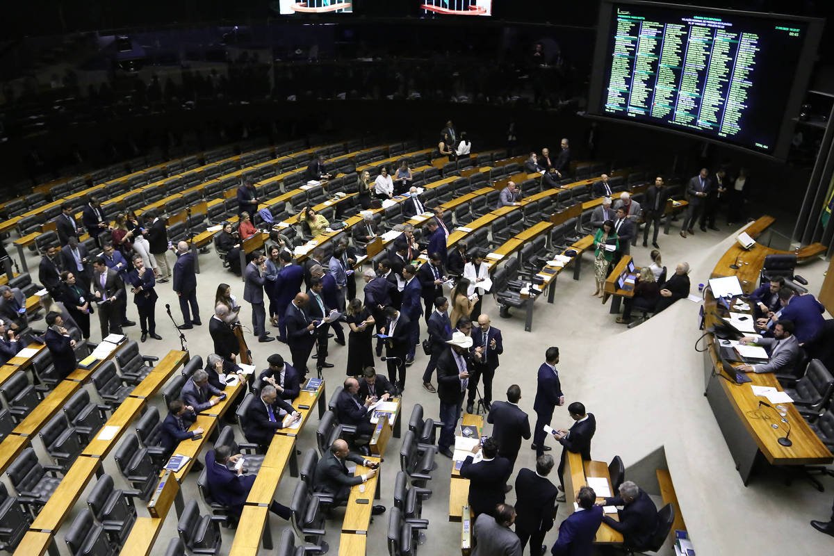 Deputados e assessores circulam e conversam entre as fileiras de bancos na Câmara dos Deputados. Painel eletrônico exibe votação em andamento. Ambiente interno com iluminação artificial e bandeiras brasileiras ao fundo.